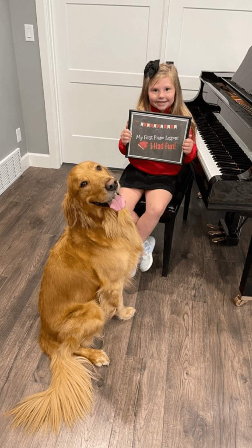 Young student at the grand piano holding a My First Piano Lesson sign, with studio dog Ollie